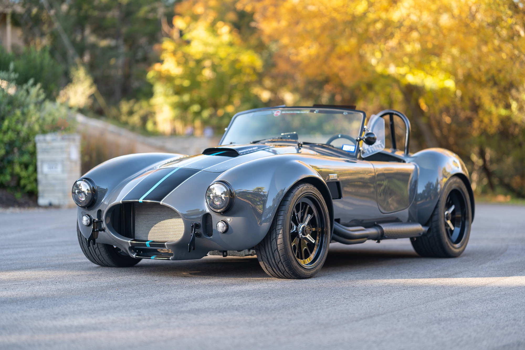Gray 1965 Backdraft Cobra with dual blue racing stripes, black rims, and side pipes.