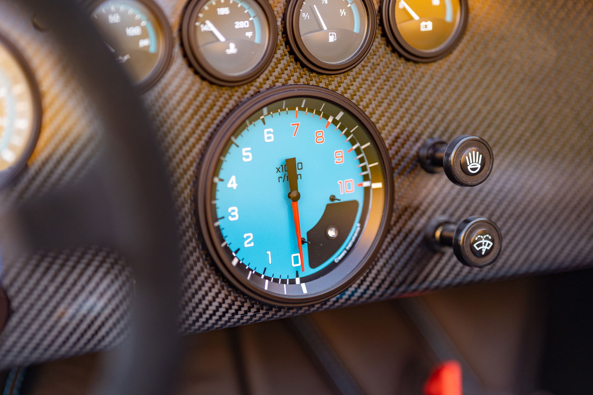 Close-up of a 1965 Backdraft Cobra's carbon fiber dash, featuring a blue tachometer and two toggle switches.