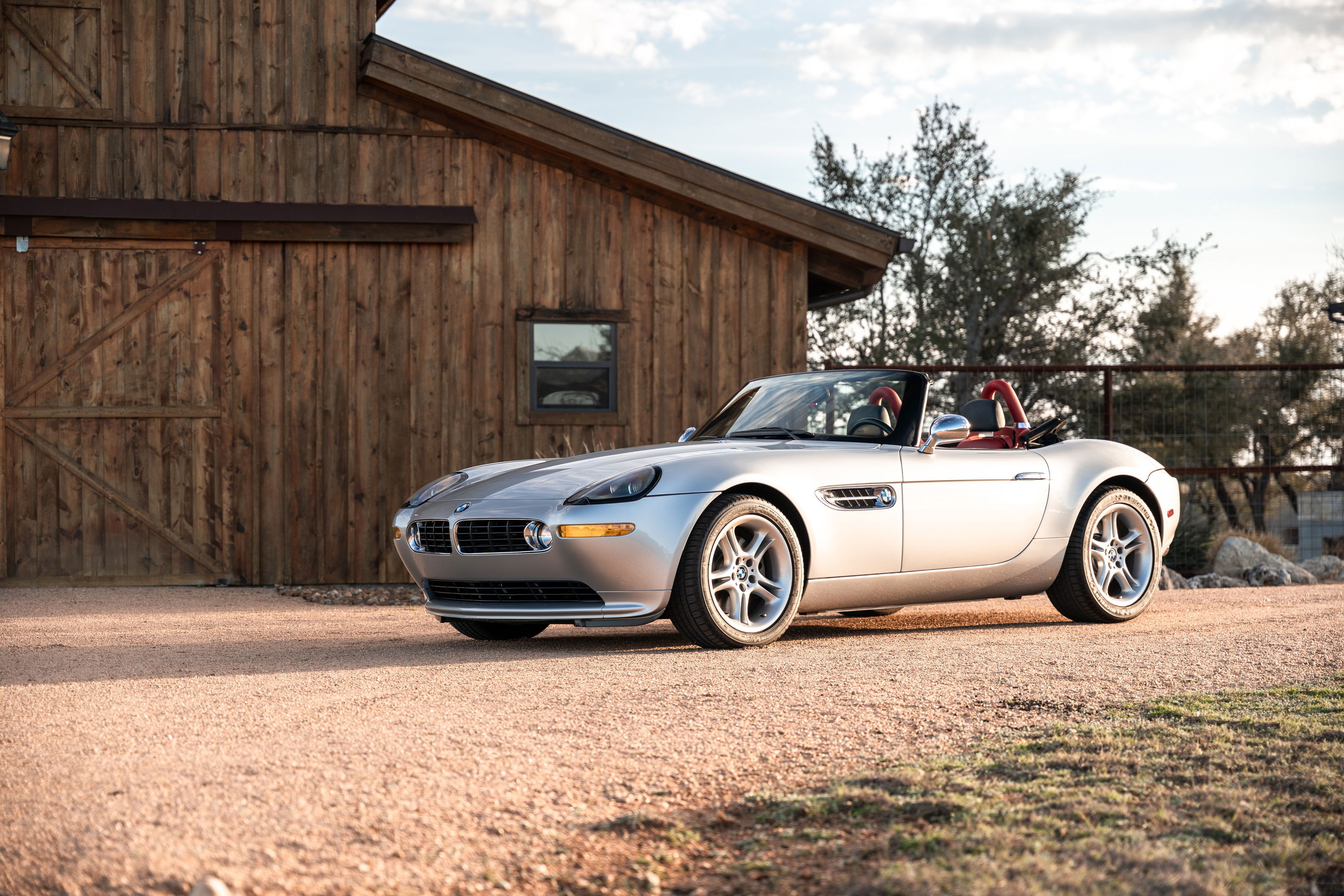 Silver 2002 BMW Z8 convertible with red interior, five-spoke rims, parked outside a wooden barn.