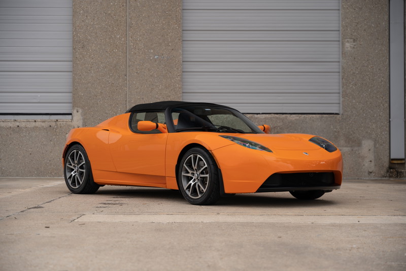 An orange 2010 Tesla Roadster with a black soft top, silver alloy rims, and a black front fascia.