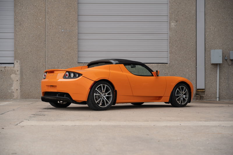 Side view of a metallic orange 2010 Tesla Roadster with dark gray multi-spoke rims.