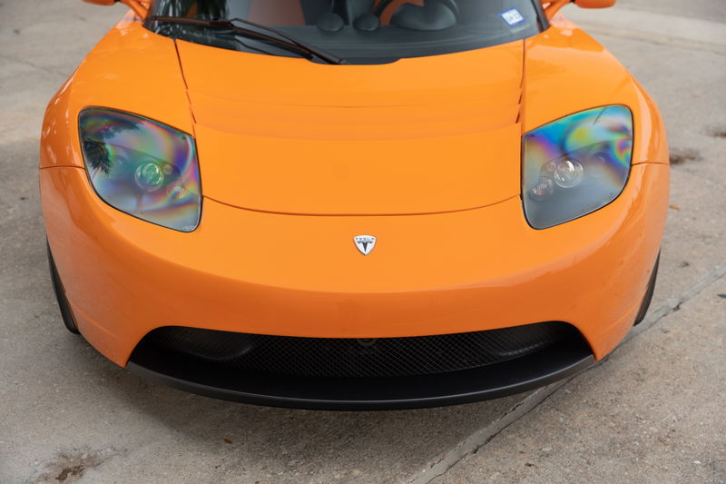 Front view of a bright orange 2010 Tesla Roadster featuring a carbon fiber lower grille and multi-colored headlight lenses.