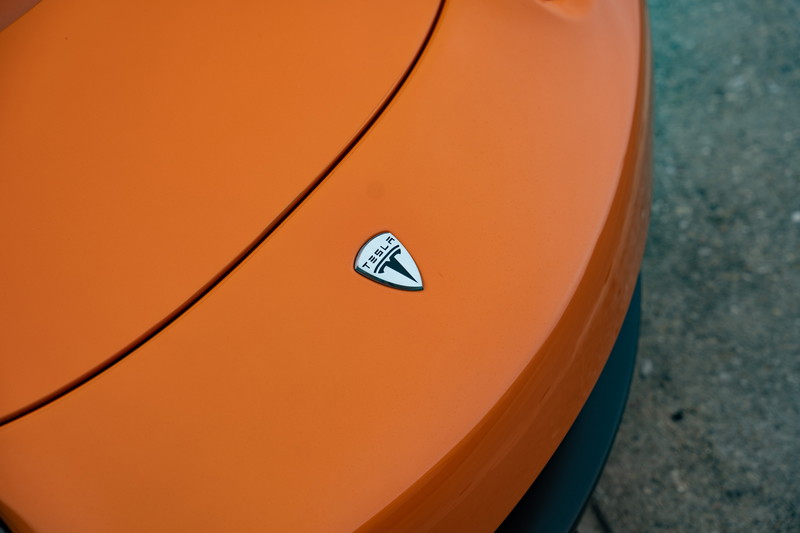 Close up of the Tesla badge on the hood of a metallic orange 2010 Tesla Roadster.