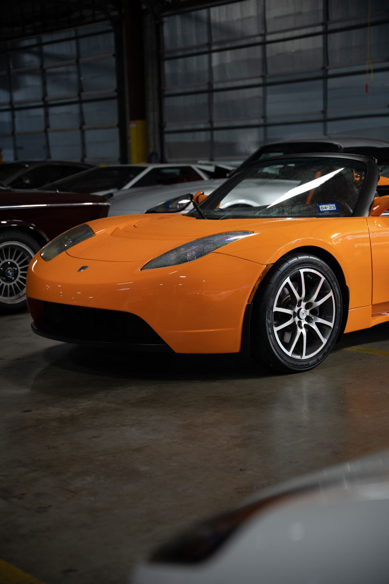 An orange 2010 Tesla Roadster sports a black front bumper and alloy rims.