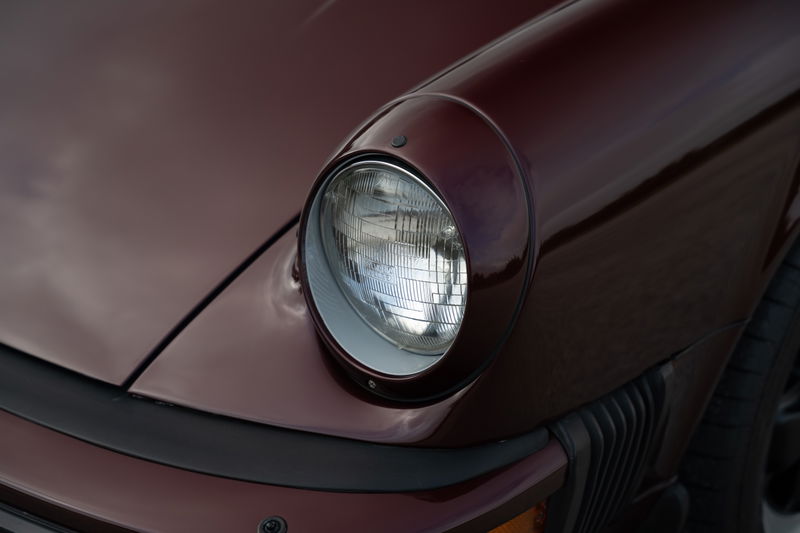 Ruby red metallic 1983 Porsche 911 SC Cabriolet headlight detail with chrome trim and black bumper.