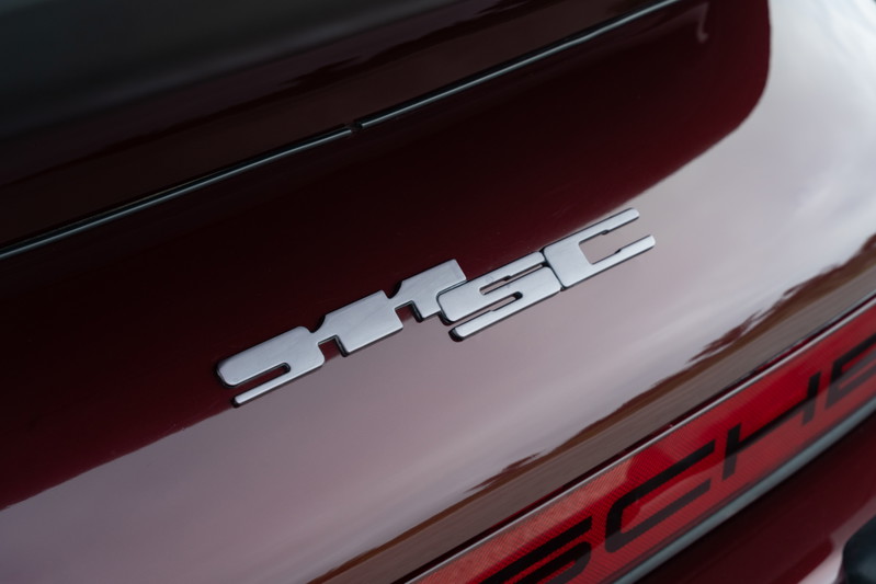 Close-up of a silver "SC" badge on the rear decklid of a ruby red 1983 Porsche 911SC Cabriolet.