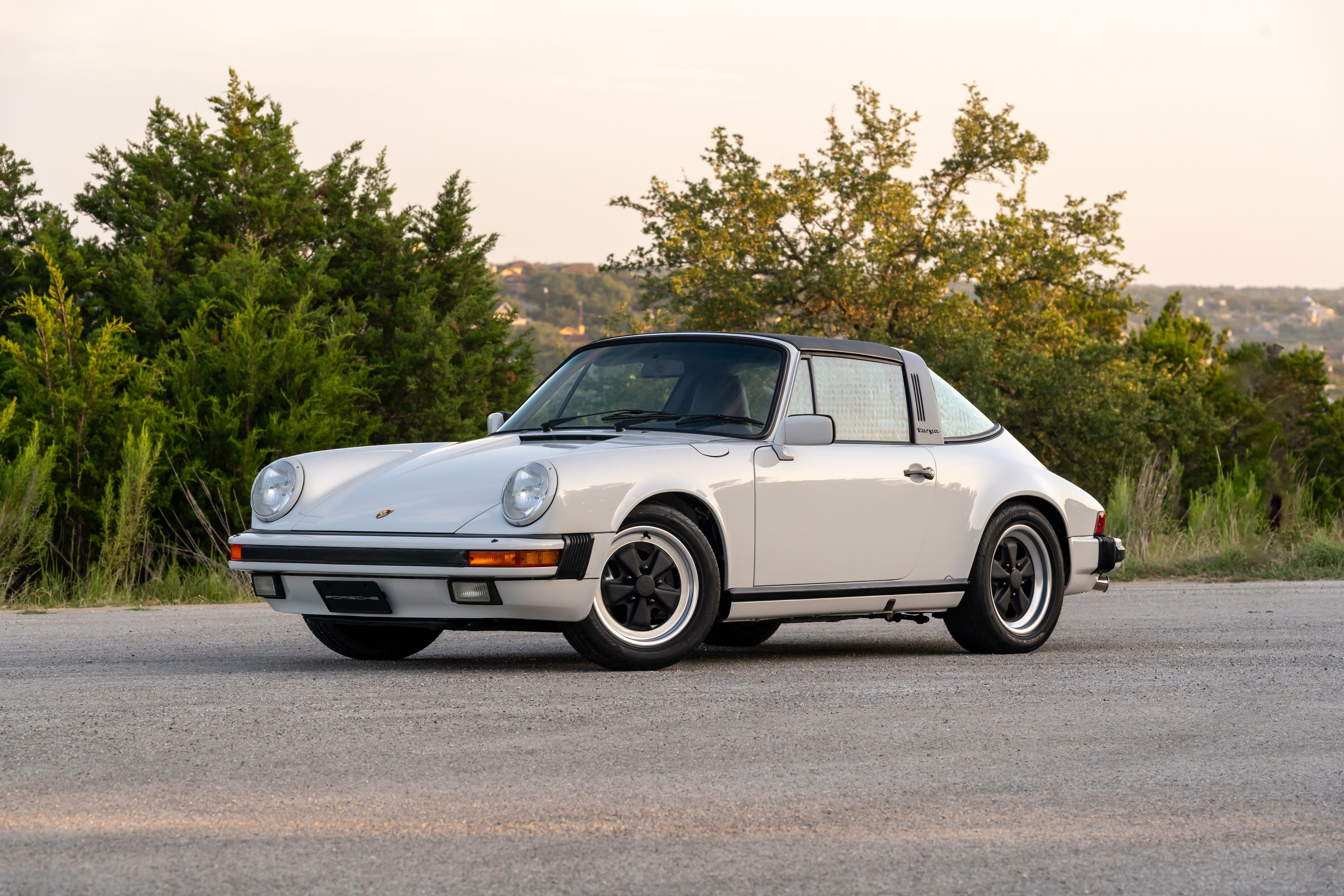 A light grey 1987 Porsche 911 Carrera Targa with black Fuchs rims and chrome trim.