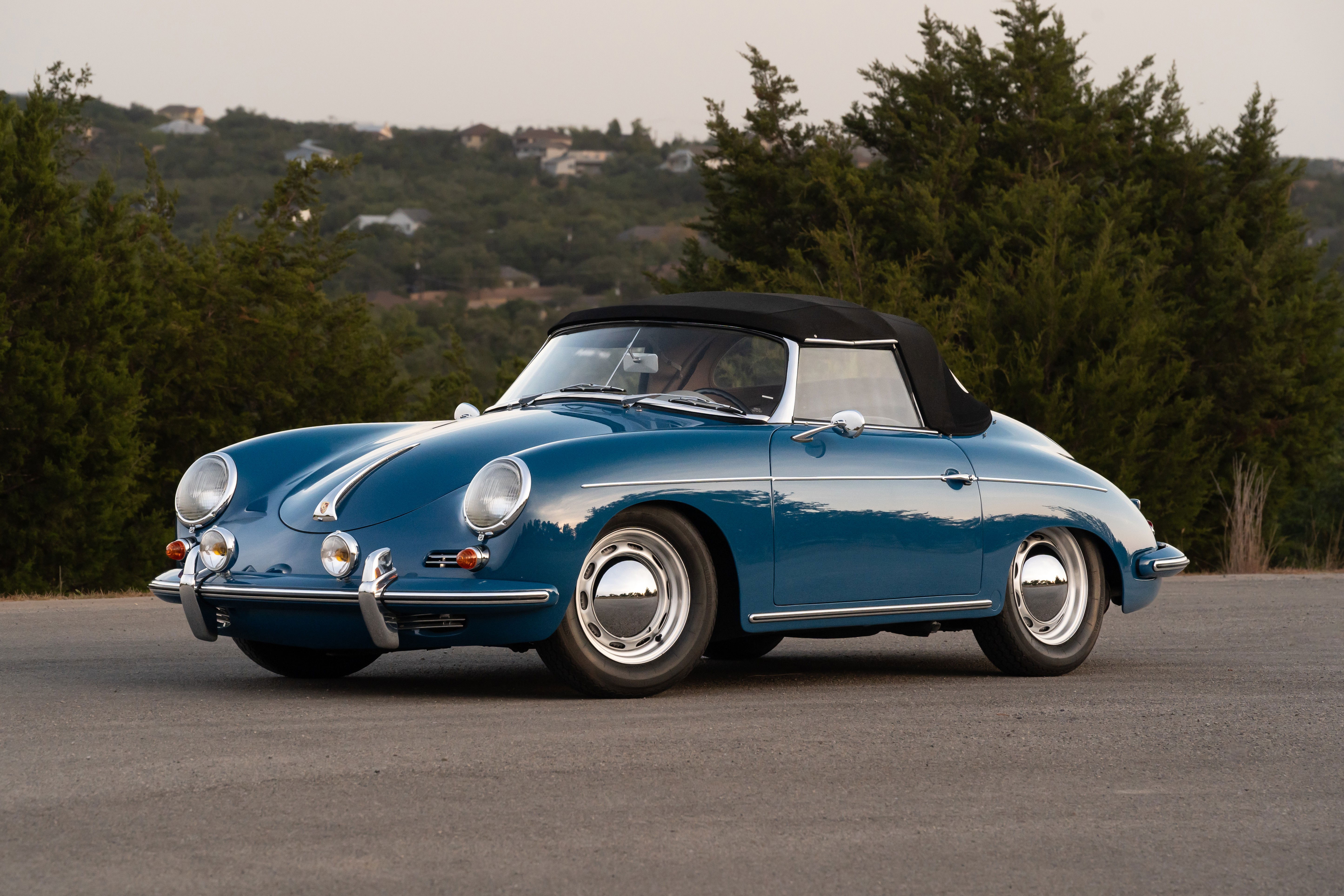 A blue 1960 Porsche 356B Roadster with chrome hubcaps and a black convertible top sits on asphalt.