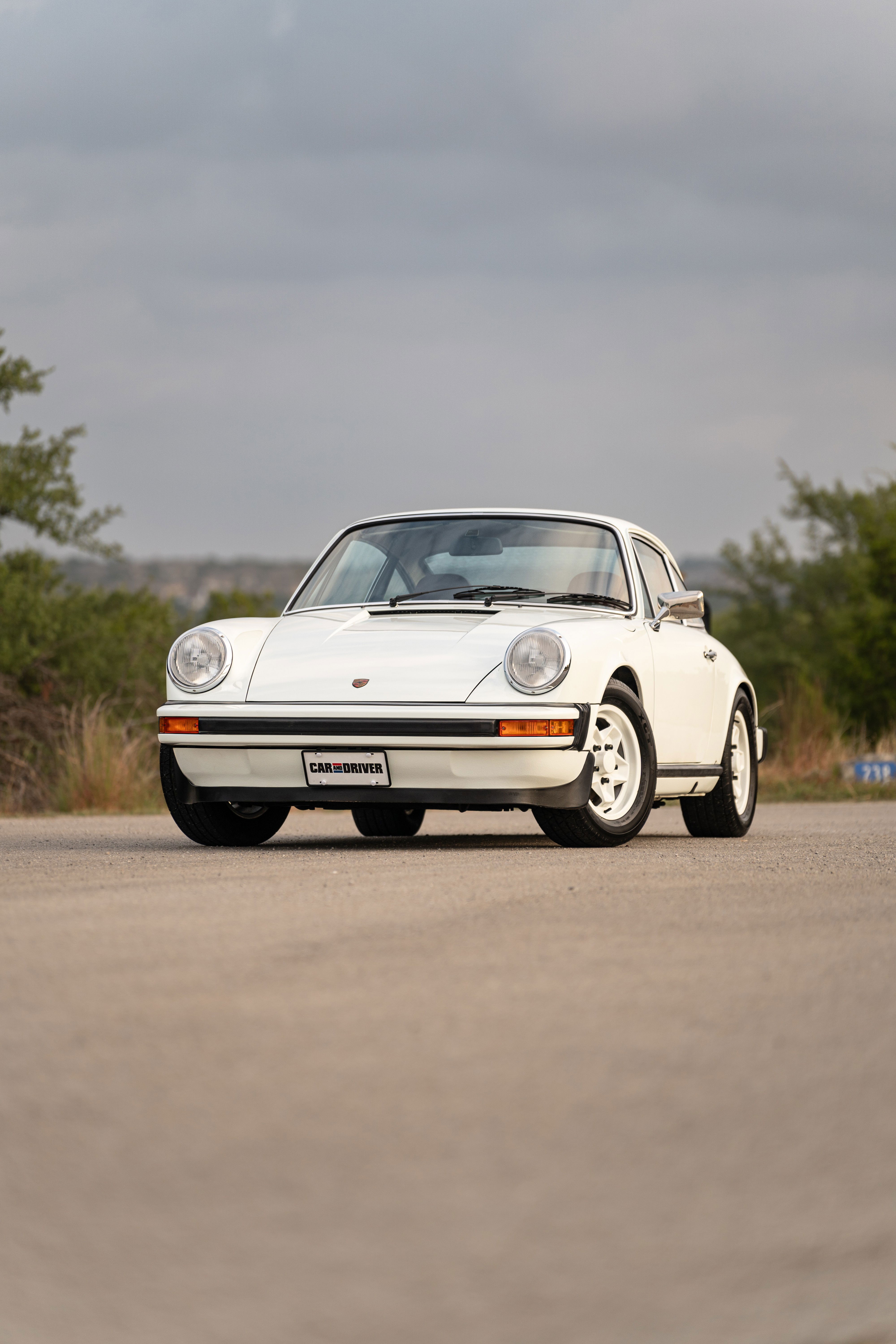 A white 1974 Porsche 911 coupe with chrome trim and Fuchs wheels sits on asphalt.
