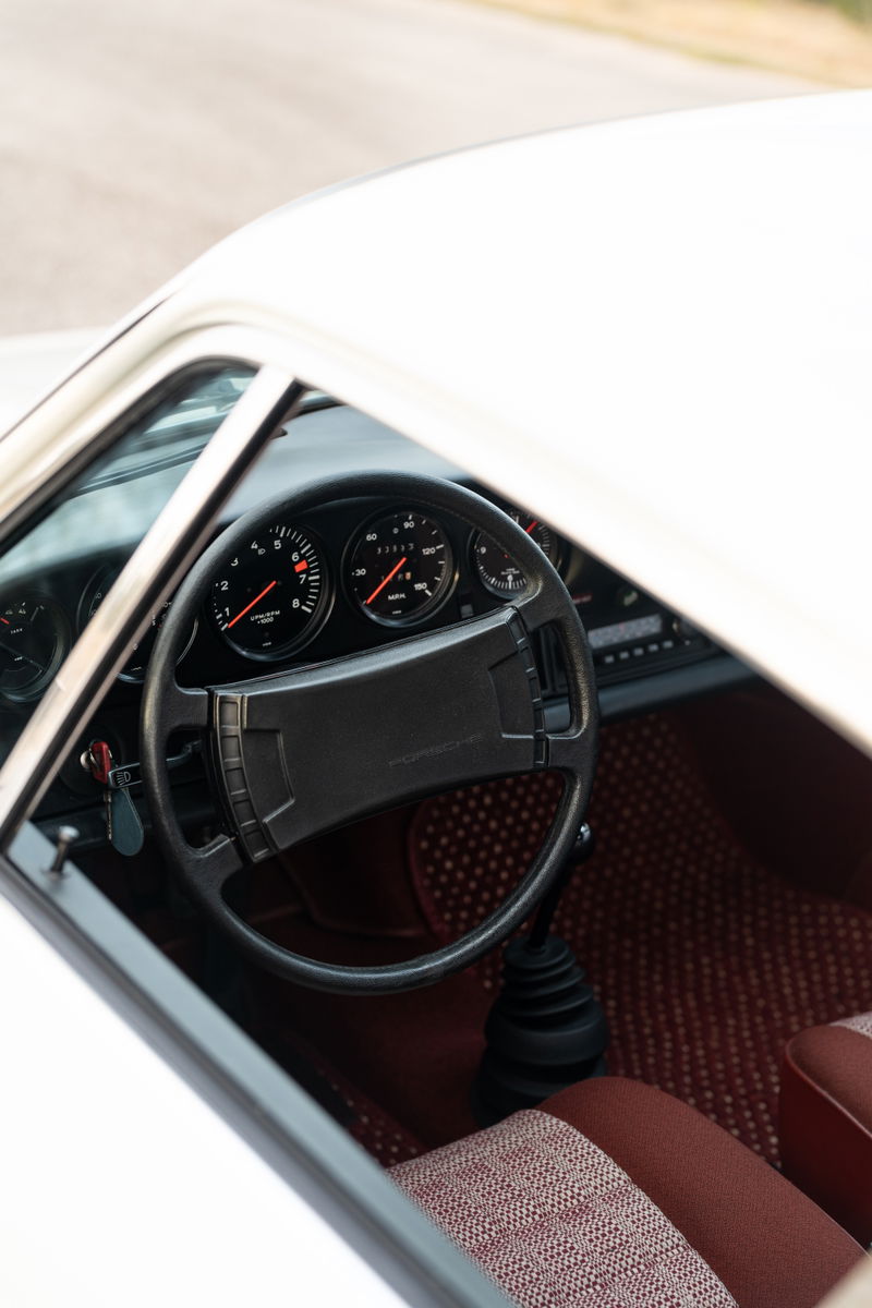 Interior of a white 1974 Porsche 911 Coupe, featuring a black steering wheel, red tartan seats, and a five-speed manual shifter.