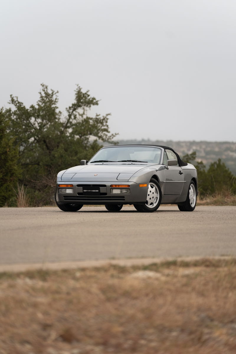 A grey 1990 Porsche 944 S2 Cabriolet with multi-spoke rims parked on a road.