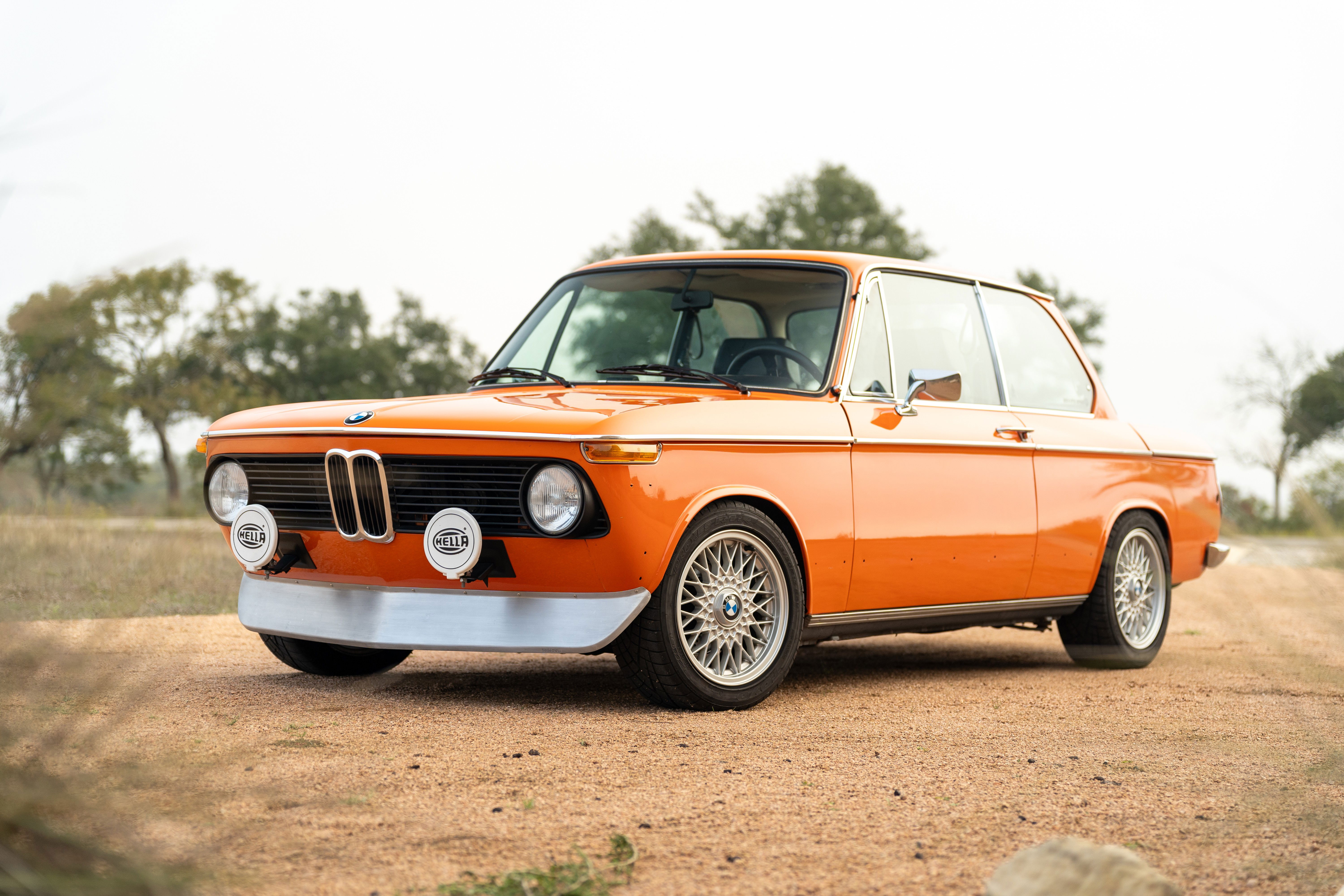 An orange 1976 BMW 2002 with classic BMW snowflake rims and Hella driving lights.