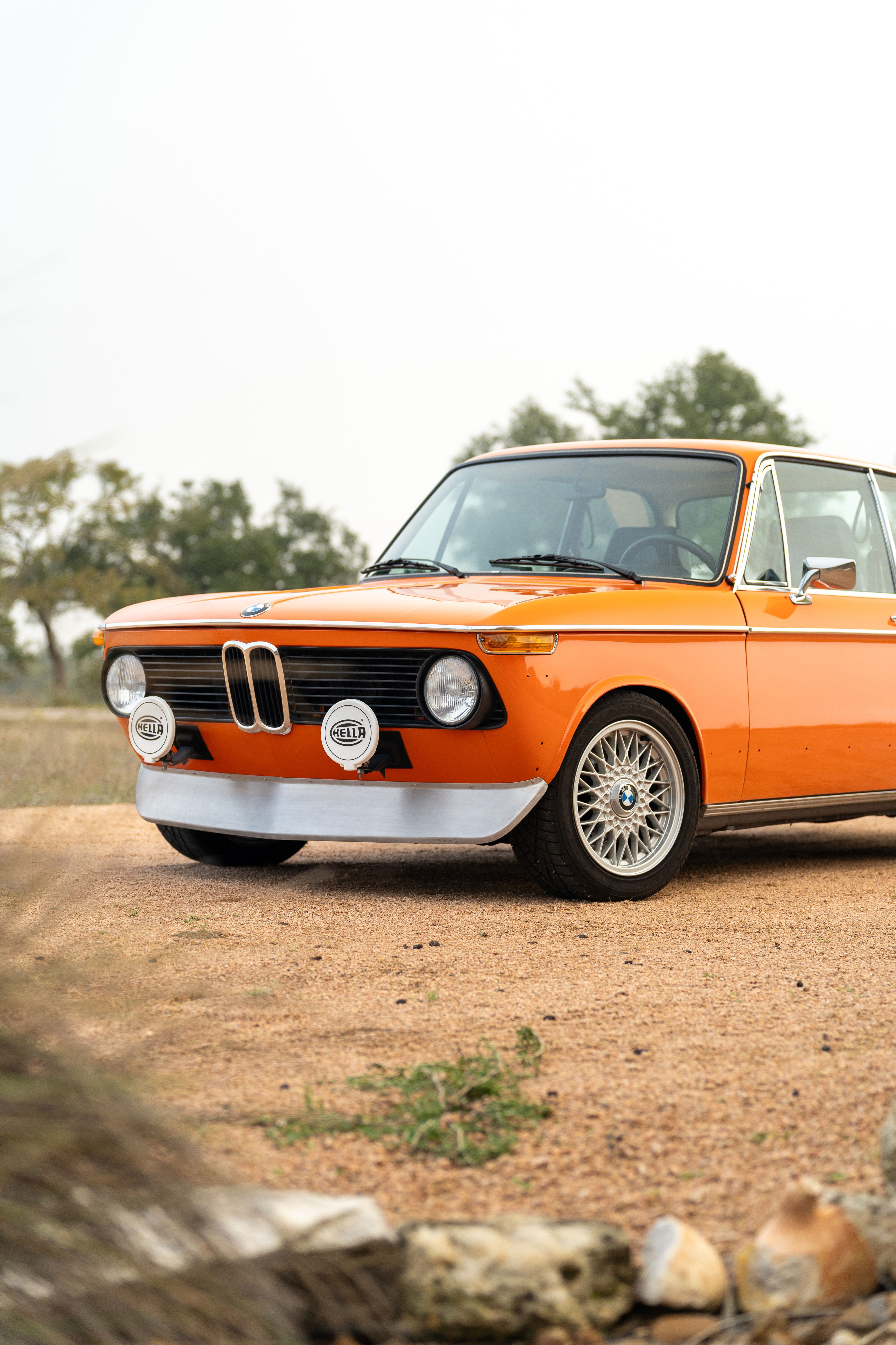 Orange 1976 BMW 2002 with silver front spoiler, Hella lights, and BBS mesh rims.