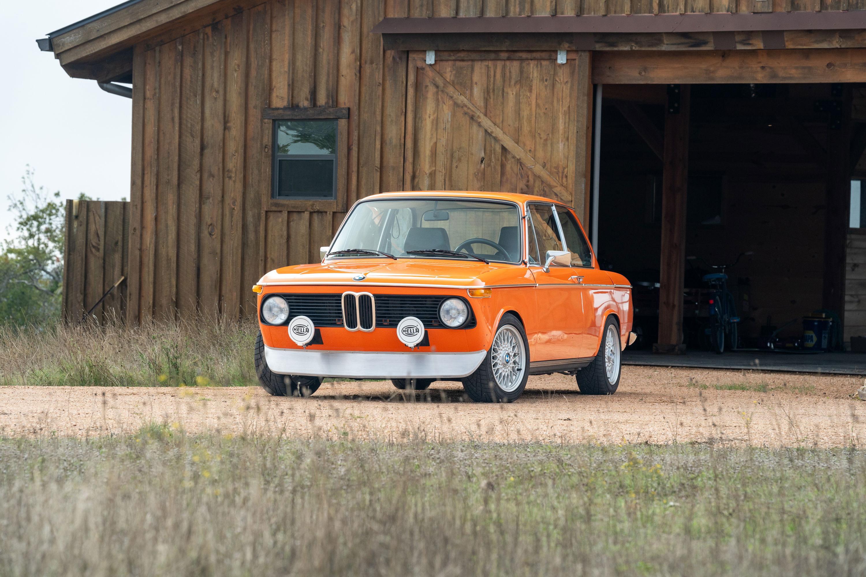 Orange 1976 BMW 2002 with vintage rally lights and silver multi-spoke rims.