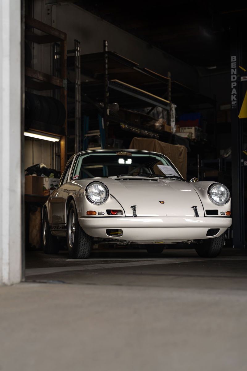 A white 1972 Porsche 911 Carrera RS 2.7 sits in a workshop, showcasing its chrome trim and classic Fuchs rims.