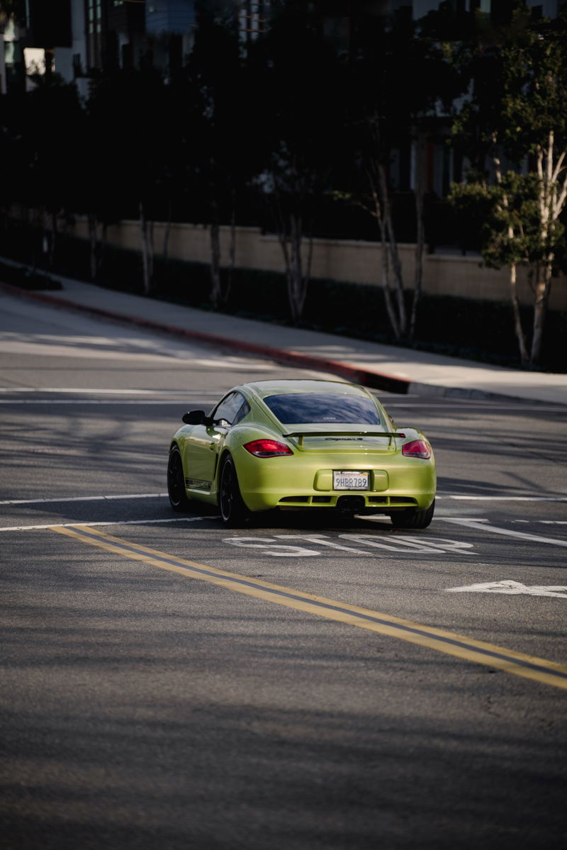 Rear view of a lime green Porsche Cayman S with black rims and a spoiler driving on the road.
