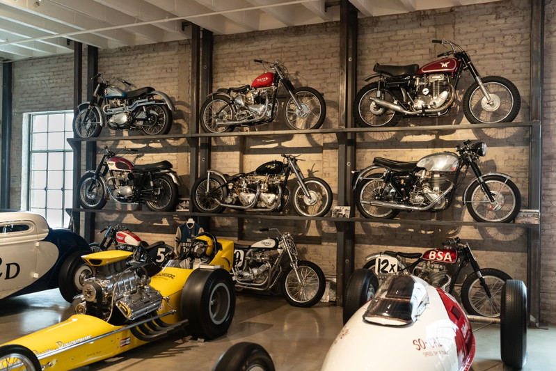 Vintage motorcycles and a yellow dragster on display shelves at the Lit Show Los Angeles 2024.