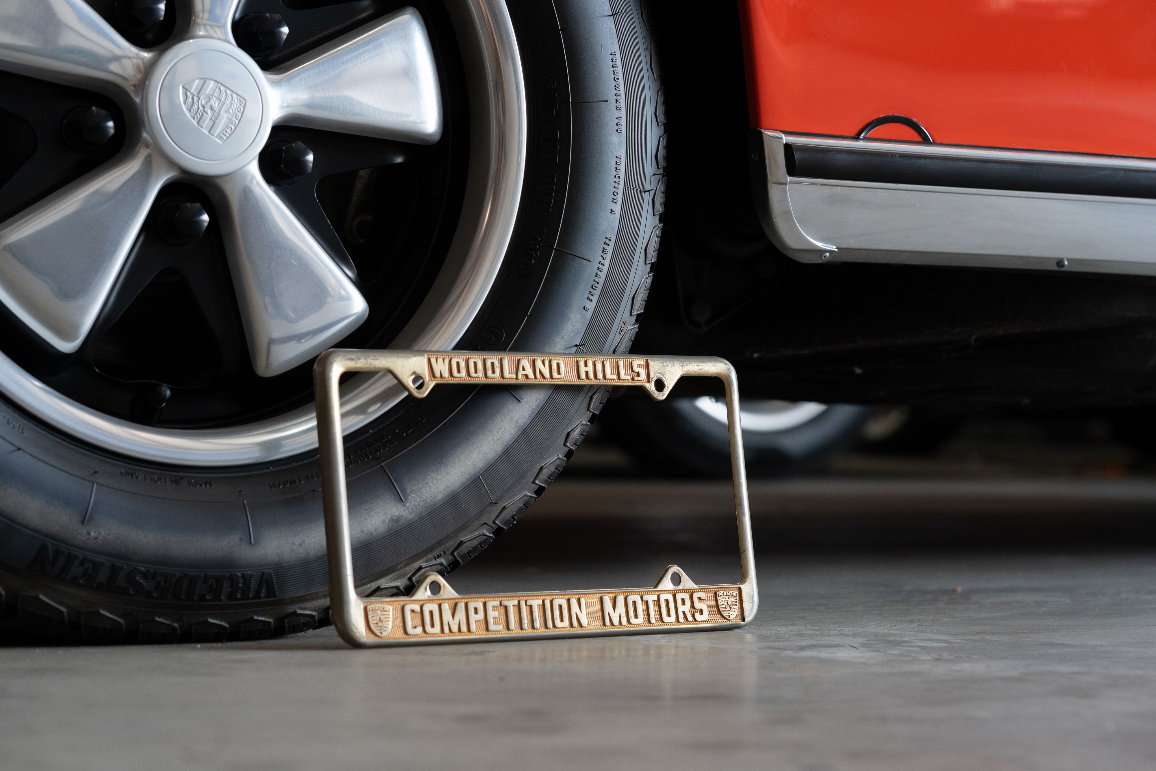 Close-up of a vintage Porsche's Fuchs rim and orange paint with a &quot;Competition Motors&quot; license plate frame.