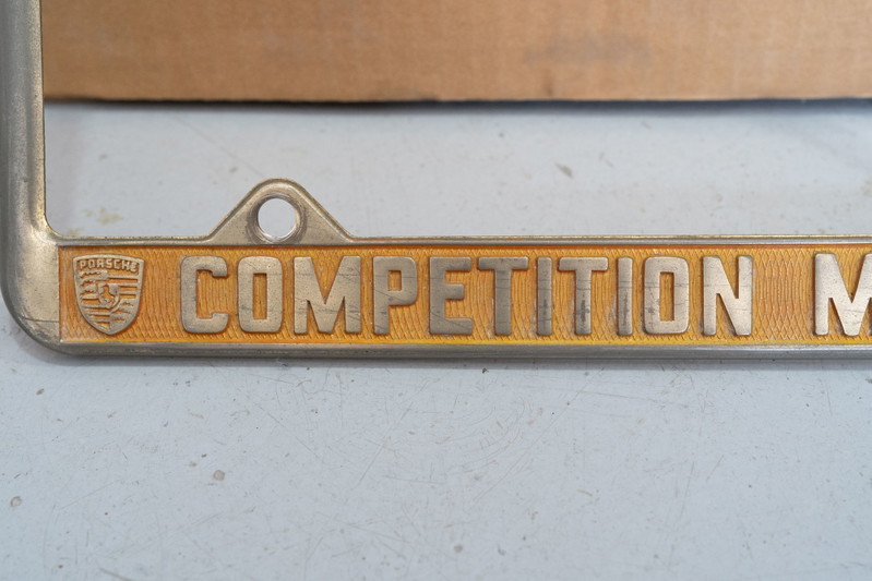 Close-up of a vintage Porsche Competition Motors license plate frame with gold trim and emblem.
