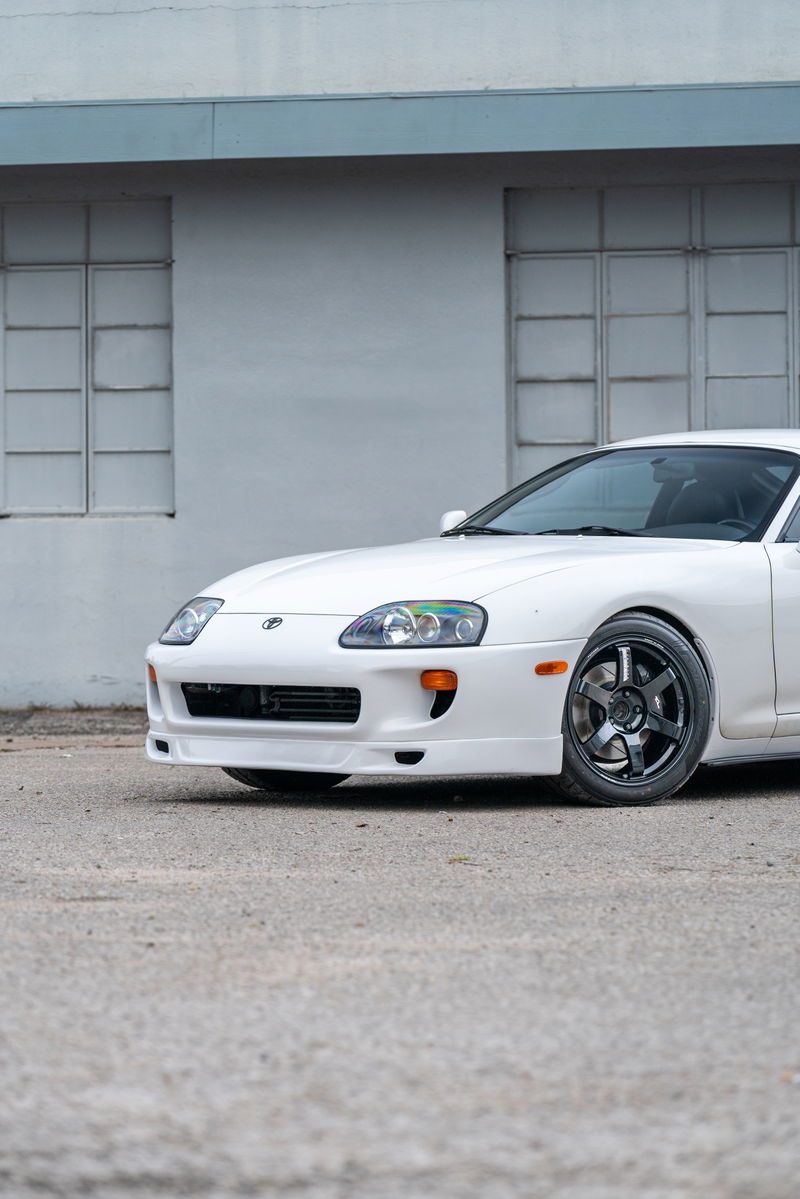 White 1994 Toyota Supra with aftermarket dark grey rims, parked on asphalt.