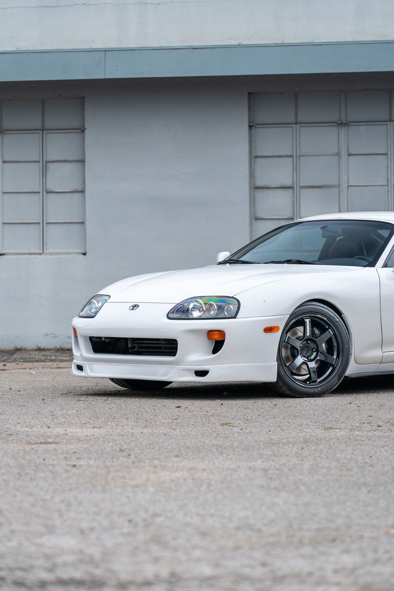 White 1994 Toyota Supra with aftermarket dark grey rims, parked on asphalt.