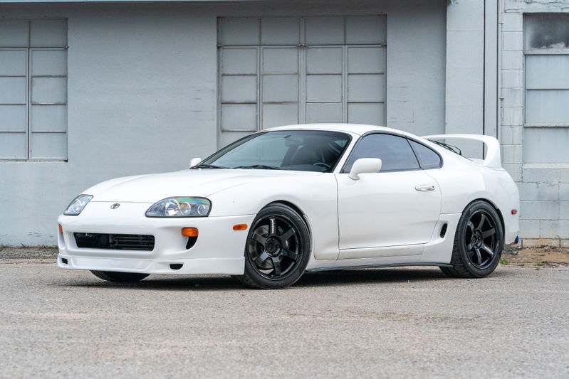 White 1994 Toyota Supra with aftermarket front bumper, rear spoiler, and black rims.