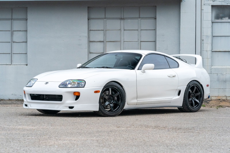 White 1994 Toyota Supra with aftermarket front bumper, rear spoiler, and black rims.