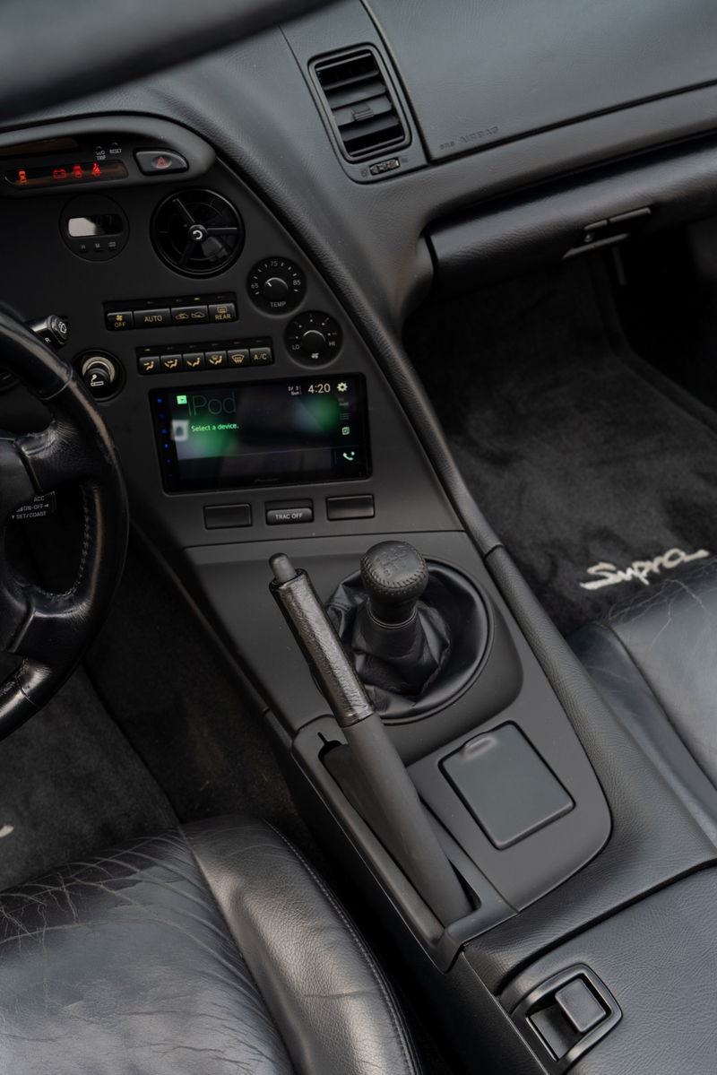 Black leather interior of a 1994 Toyota Supra, featuring a 5-speed manual shifter, modern infotainment screen, and "Supra" floor mat.