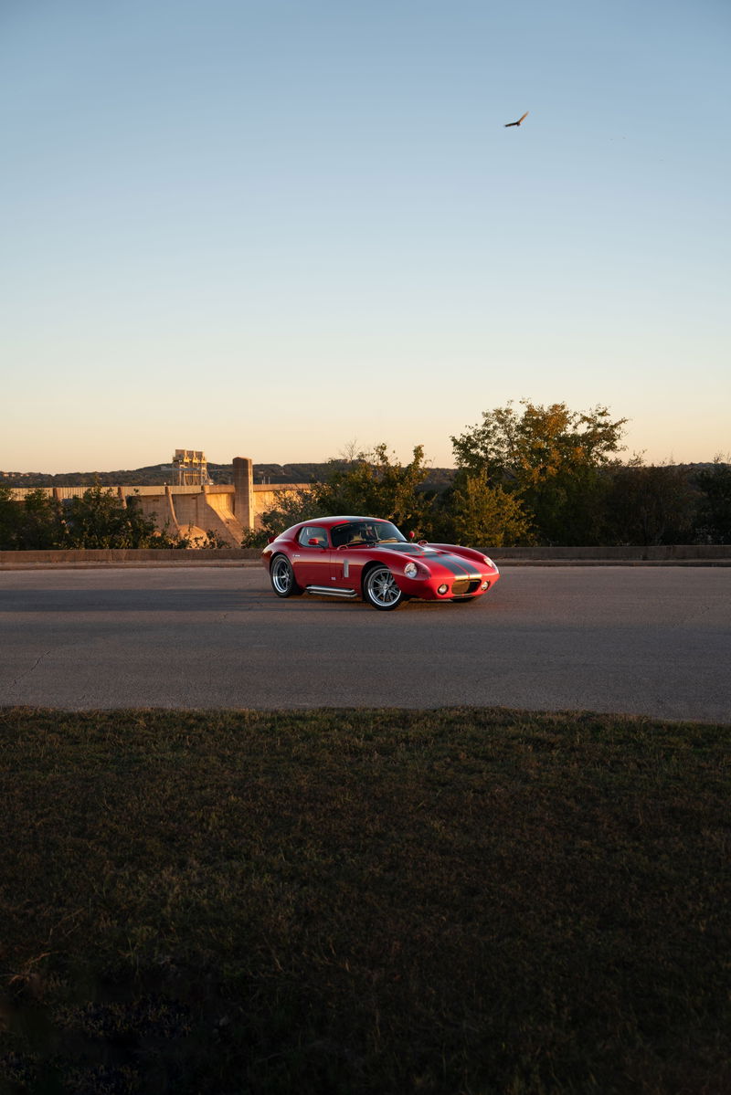Red 1965 Superformance Shelby Daytona CSX with black racing stripes and chrome rims drives past a dam.