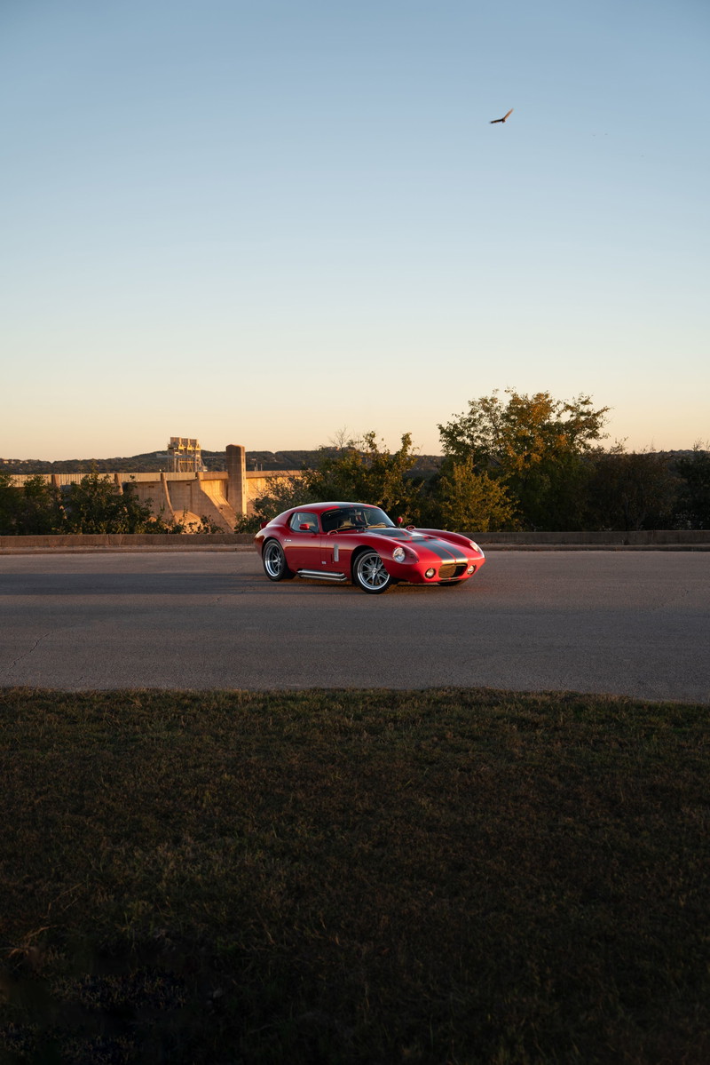Red 1965 Superformance Shelby Daytona CSX with black racing stripes and chrome rims drives past a dam.
