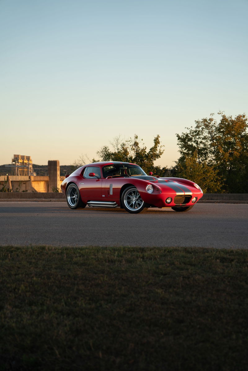 A red 1965 Superformance Shelby Daytona CSX with dual racing stripes on the hood, and chrome side pipes.