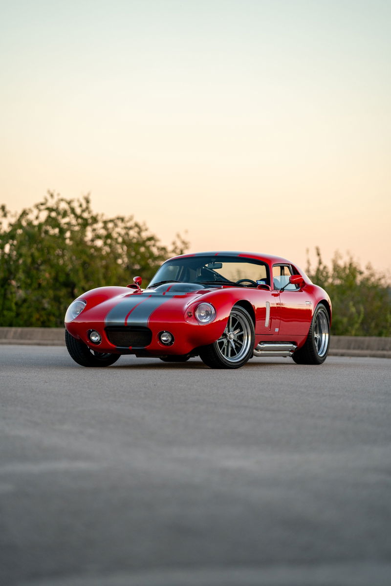 Red 1965 Superformance Shelby Daytona coupe with grey racing stripes and chrome rims.