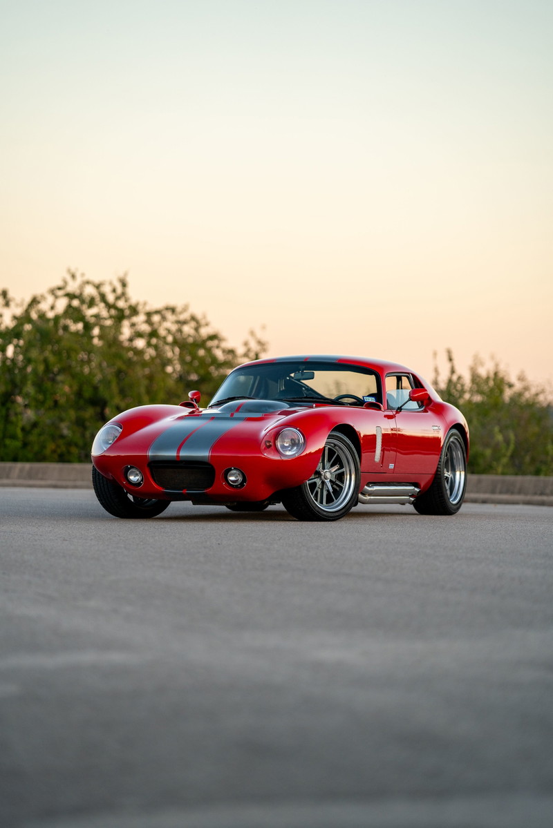 Red 1965 Superformance Shelby Daytona coupe with grey racing stripes and chrome rims.