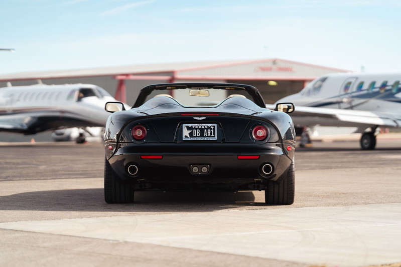 Rear view of a black 2003 Aston Martin DB AR1 Zagato convertible, parked on tarmac by private jets.