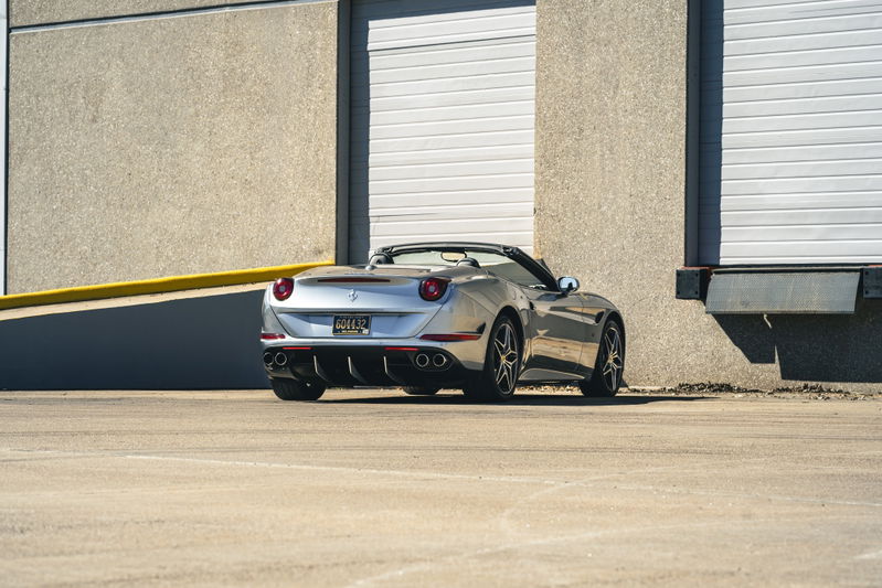 Silver 2016 Ferrari California T convertible with dark multi-spoke rims parked by a loading dock.