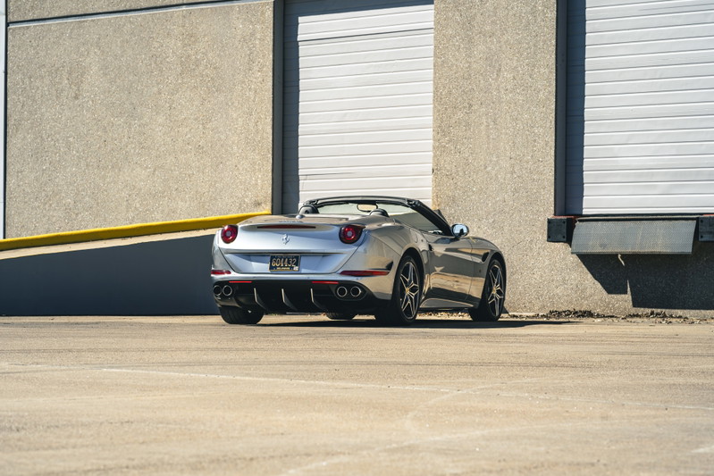 Silver 2016 Ferrari California T convertible with dark multi-spoke rims parked by a loading dock.