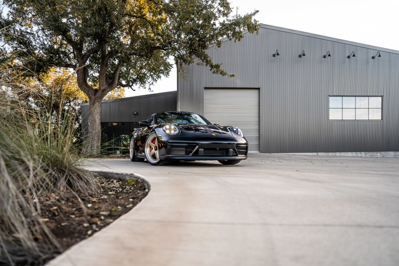 Black 2023 Porsche 911 Carrera 4 GTS with striking copper rims, parked in front of a modern warehouse.