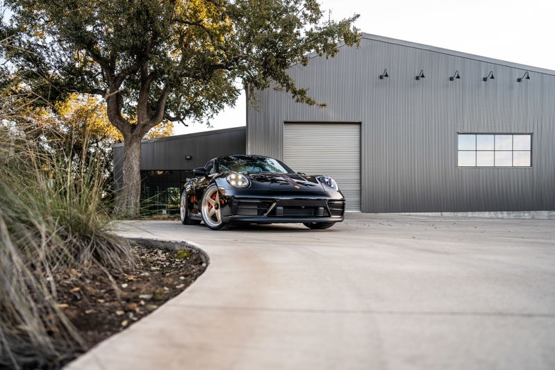 Black 2023 Porsche 911 Carrera 4 GTS with striking copper rims, parked in front of a modern warehouse.