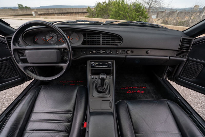 Interior view of a black 1986 Porsche 944 Turbo with leather seats and red "Porsche Turbo" floor mats.