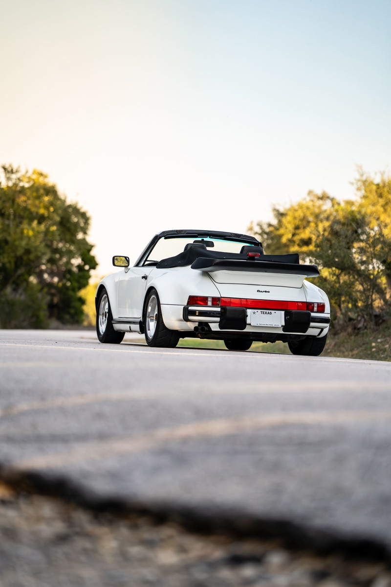 White 1988 Porsche 930 Turbo slantnose convertible with black soft top and whale tail spoiler.