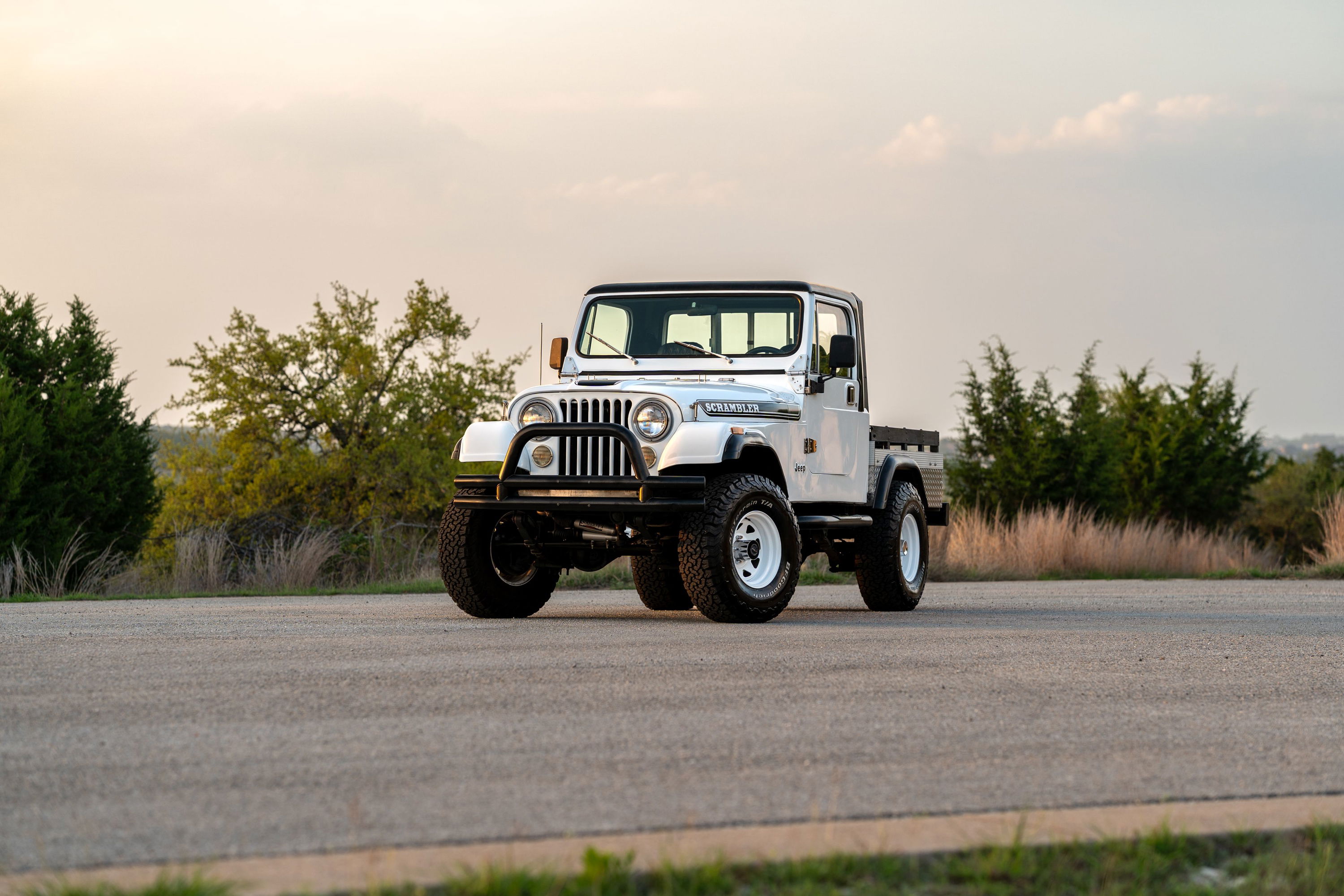 White 1983 Jeep Scrambler pickup with white rims and off-road tires parked on asphalt.