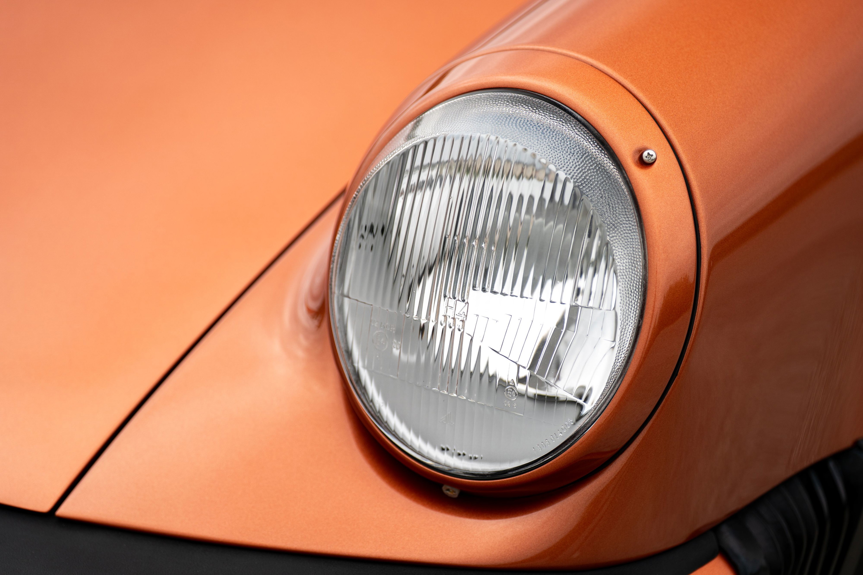 Close-up of a 1986 Porsche 930 Turbo's round headlight in orange metallic paint.