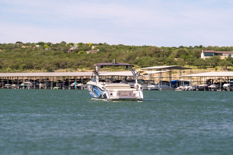 Rear view of a white Cobalt R7 Surf boat with blue livery, cruising on a lake near a marina.