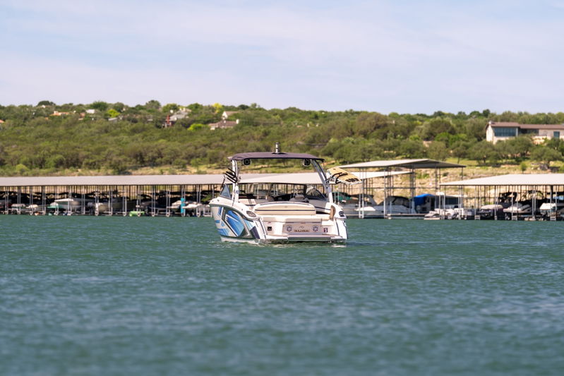 Rear view of a white Cobalt R7 Surf boat with blue livery, cruising on a lake near a marina.