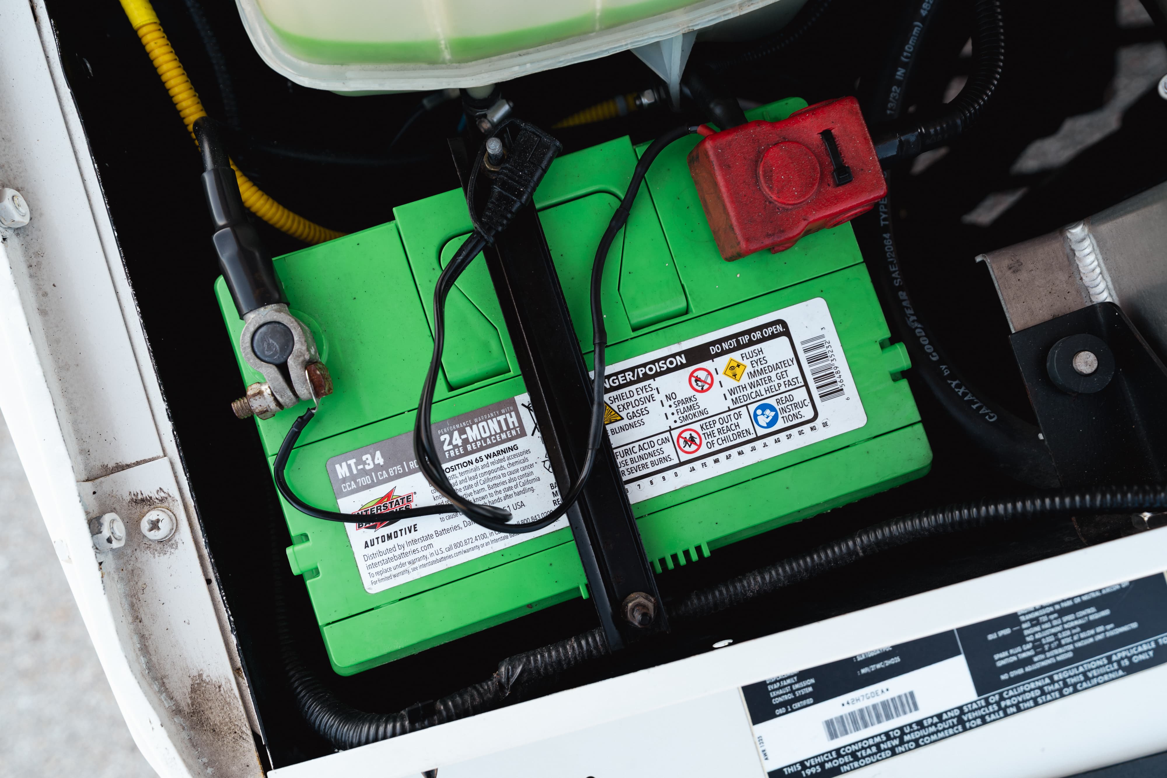 Green automotive battery (MT-34) with terminals and red fuse cover in engine bay of white 1995 Land Rover Range Rover County.