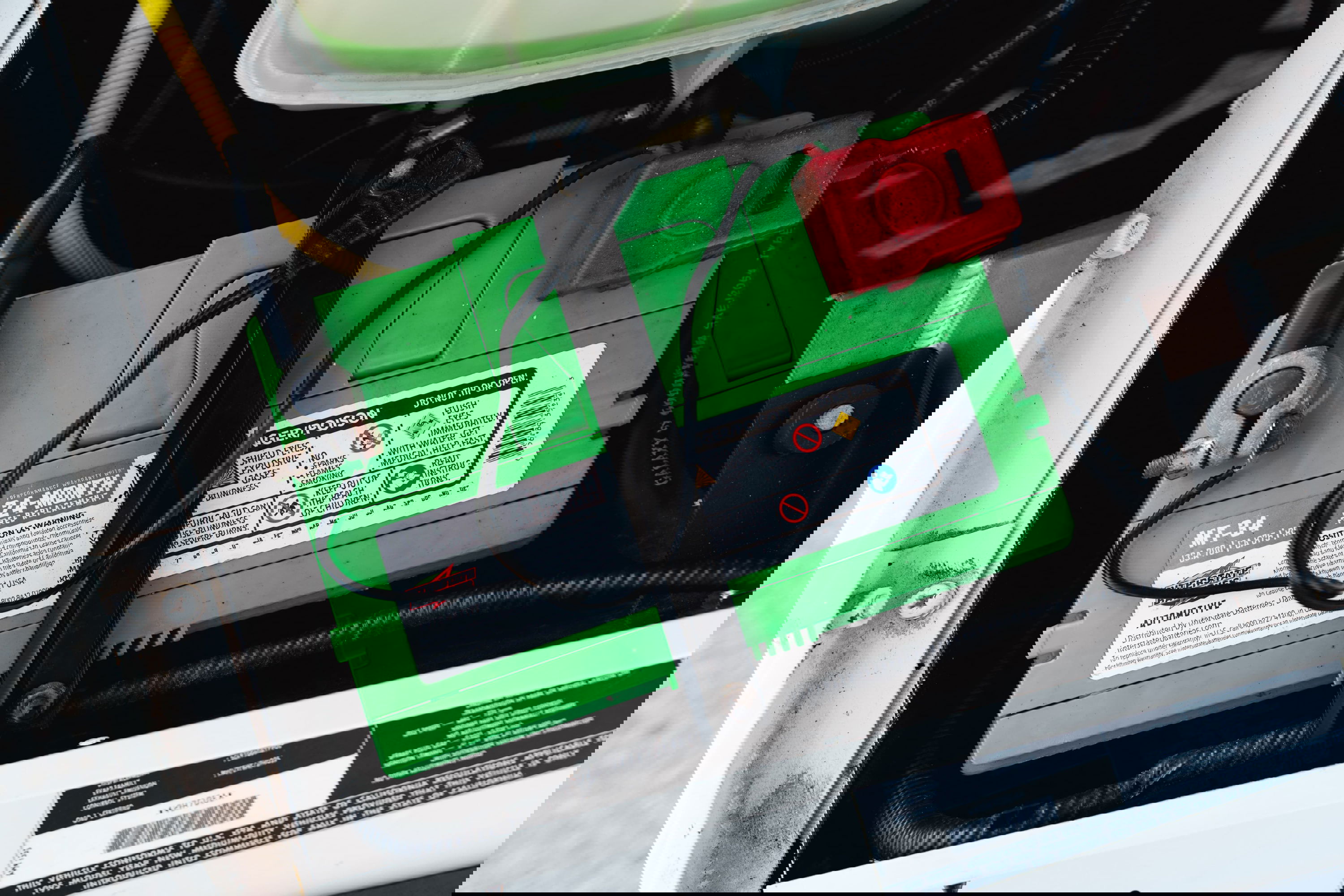 Green automotive battery (MT-34) with terminals and red fuse cover in engine bay of white 1995 Land Rover Range Rover County.