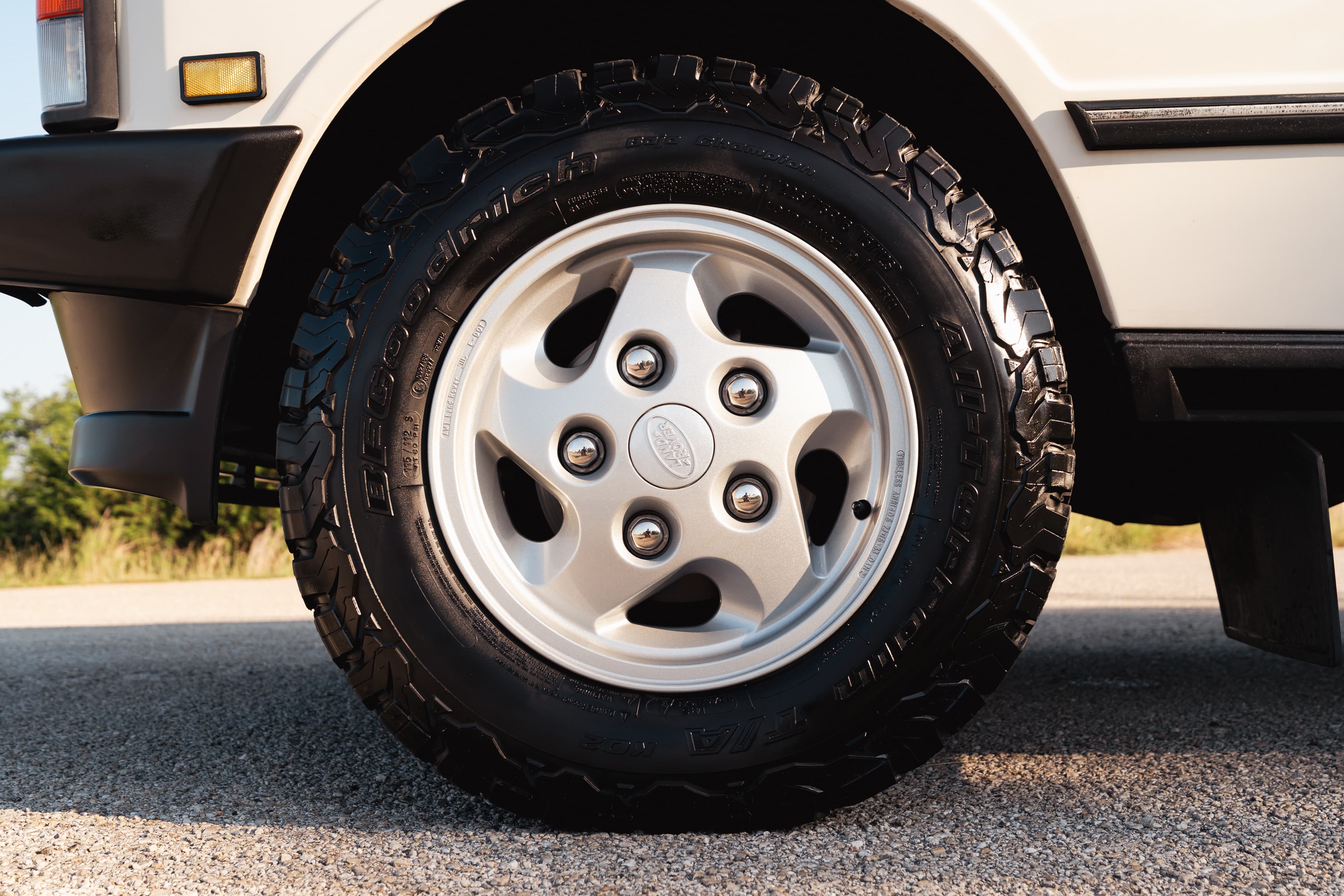 Close-up of a white 1995 Land Rover Range Rover County's silver 5-spoke rim and all-terrain tire.