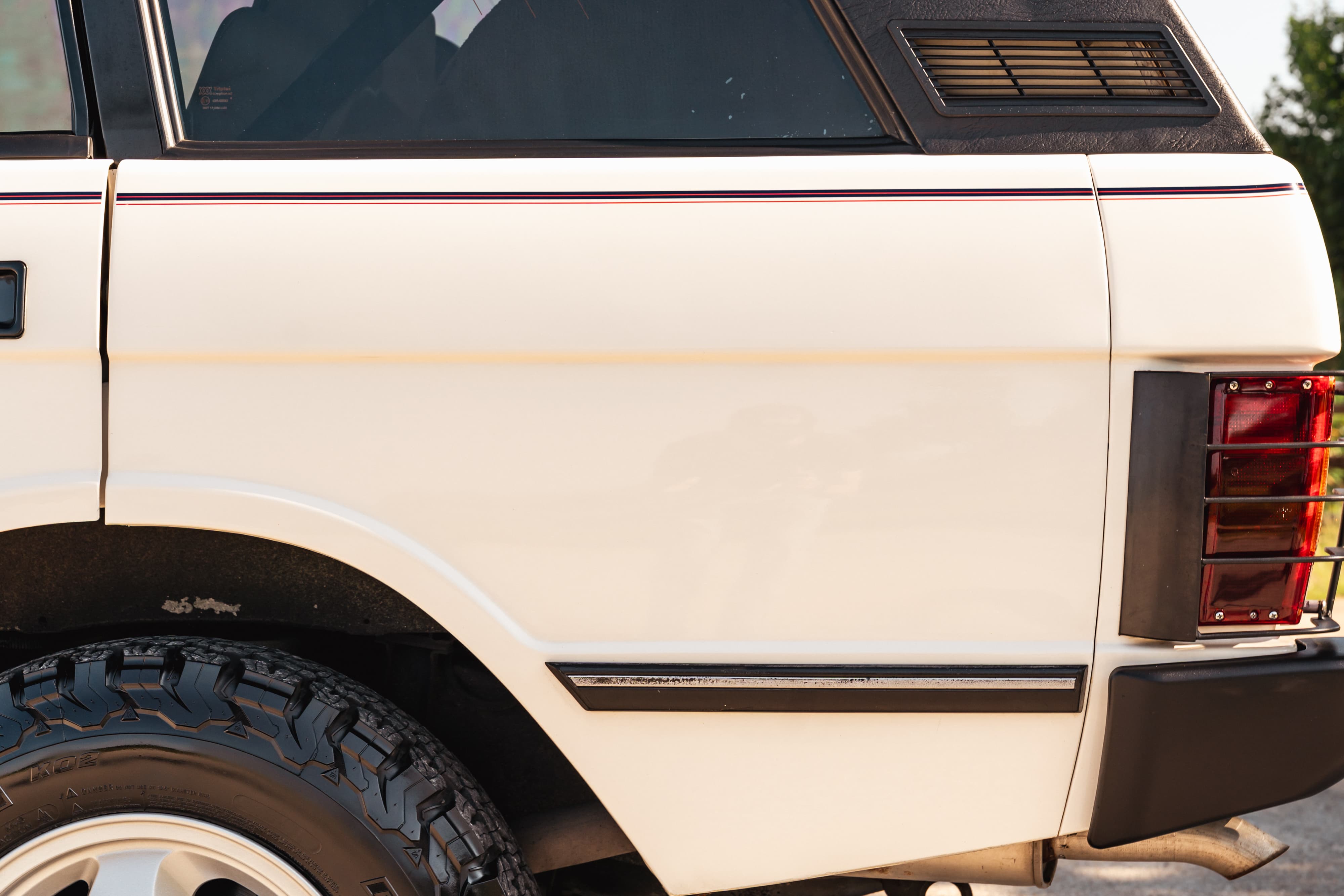 Close-up on the rear quarter panel of a white 1995 Land Rover Range Rover County. Features include a black vinyl roof, red taillight assembly, and off-road tires.