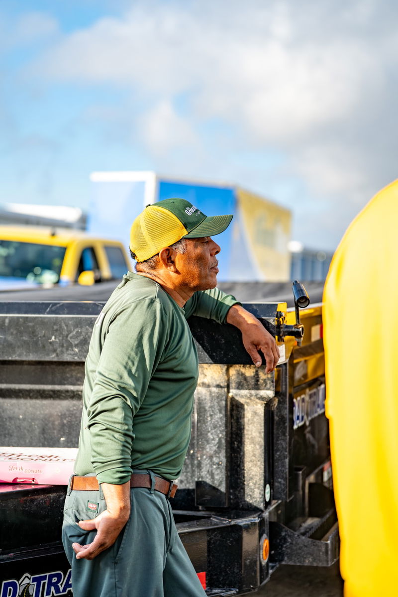 Man in green shirt & cap leans on a black utility trailer, with a yellow pickup truck in the background.