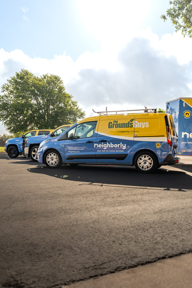 Blue Ford Transit Connect van with yellow "The Grounds Guys" livery and silver rims parked next to a blue pickup truck.